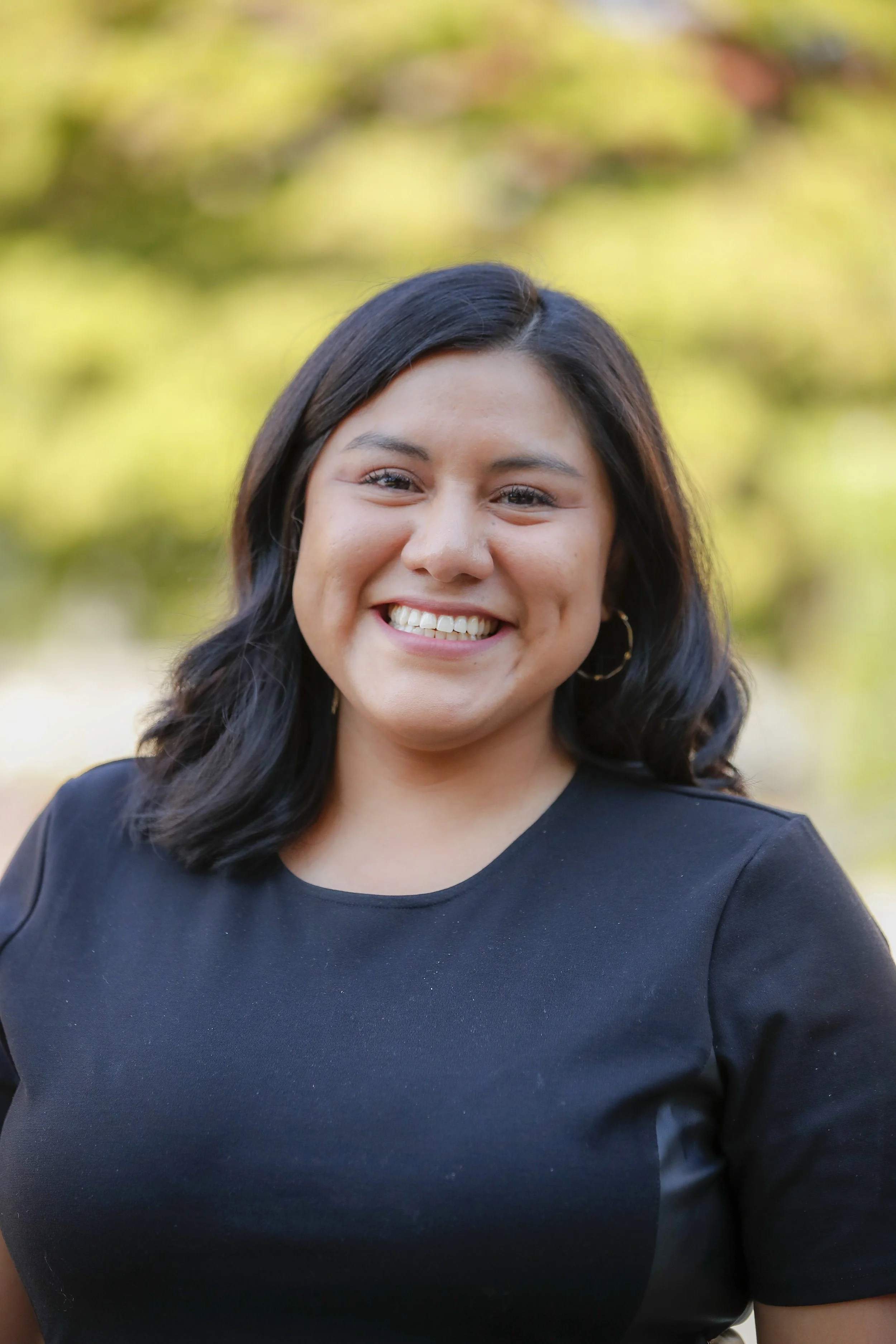 A young woman with black hair and hoop earrings smiling outdoors.
