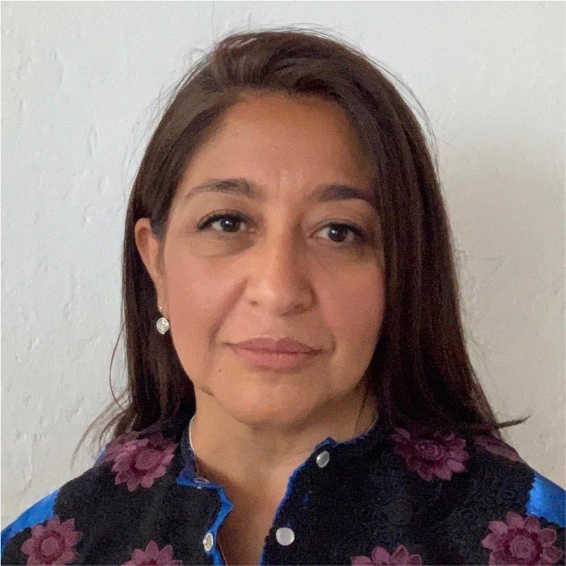 Portrait of a woman with brown hair, wearing pearl earrings and a dark blouse with a floral pattern, standing against a white wall.
