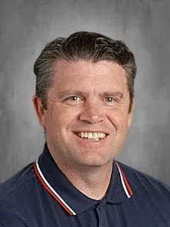 Smiling man with short, dark hair wearing a dark polo shirt with a red, white, and blue collar, in front of a gray background.