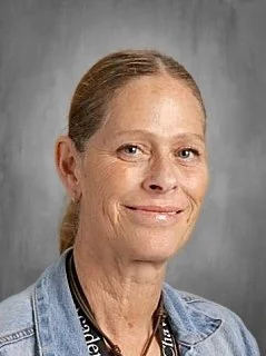 A woman with light brown hair pulled back, wearing a denim jacket and a black lanyard, smiling against a gray background.