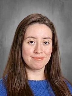 A young woman with long brown hair wearing a blue sweater, smiling subtly at the camera, against a gray background.