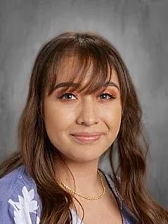 A woman with long brown hair and bangs, smiling, wearing a purple top and gold necklace, against a gray background.