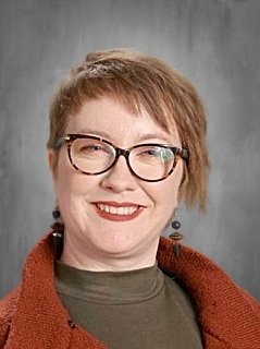 Portrait of a woman with short, light brown hair, wearing glasses, a green top, and a reddish-brown cardigan, smiling against a gray background.