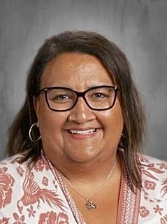A woman with glasses smiling, wearing earrings, a necklace, and a patterned top, in front of a plain background.