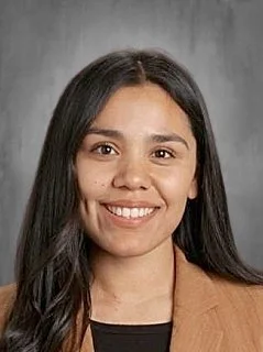 A young woman with long dark hair smiling at the camera, wearing a brown blazer and black top, against a neutral gray background.