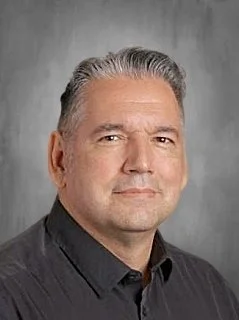 Close-up portrait of a middle-aged man with gray hair, wearing a black collared shirt, looking slightly to the right with a neutral expression against a gray background.
