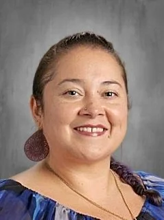 A young woman with light skin and brown hair tied back, smiling, wearing a blue top with a textured earring, standing against a gray background.