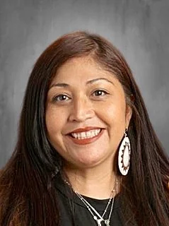 Portrait of a woman with long dark hair, smiling, wearing earrings and a necklace, against a gray background.