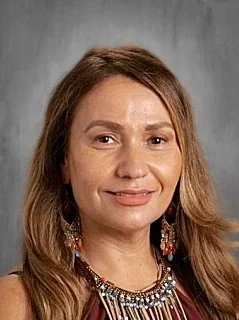 Close-up portrait of a woman with light brown hair, smiling, wearing earrings and a necklace, against a gray background.