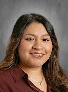 Portrait of a woman with long dark hair smiling, wearing a maroon top and a gold necklace, against a gray background.