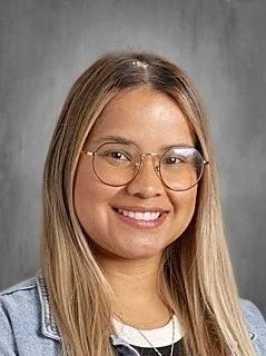 A young woman with long blonde hair, glasses, and a denim jacket, smiling at the camera against a plain gray background.