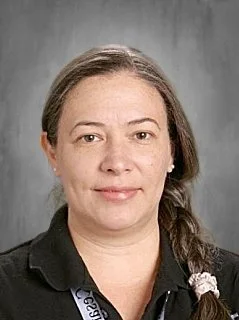 A woman with light skin and brown hair tied in a braid, wearing a black collared shirt, standing against a gray background.