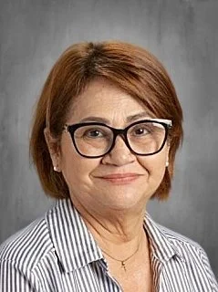A woman with short reddish-brown hair, glasses, and a striped shirt smiling at the camera against a gray background.