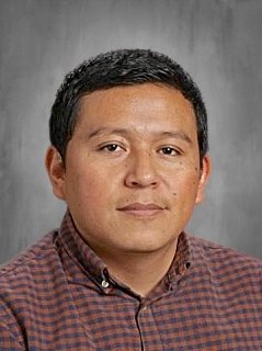 Portrait of a man with short black hair, wearing a checkered shirt, against a gray background.