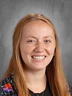Smiling young woman with long red hair, wearing a dark floral top, posed against a plain gray background.