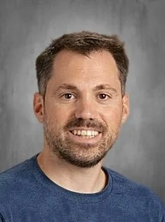 A portrait of a man with short brown hair and a beard, smiling, wearing a blue sweater against a gray background.