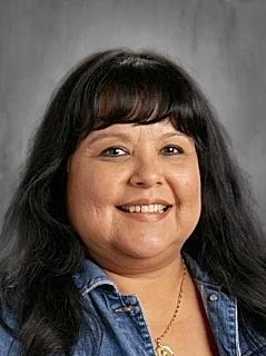 A woman with shoulder-length dark hair, smiling, wearing a denim jacket and a necklace, against a gray background.