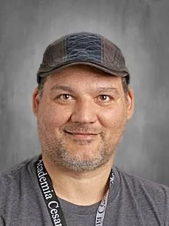 A man with a beard wearing a flat cap and a gray shirt, smiling at the camera, against a plain gray background.