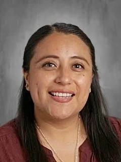 Portrait of a woman with long dark hair, smiling, wearing a burgundy top and a necklace, against a gray background.