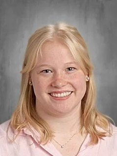 A young girl with blonde hair, wearing a light pink top, smiling against a gray background.