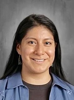 Portrait of a woman with long black hair wearing earrings and a blue uniform shirt, smiling against a gray background.