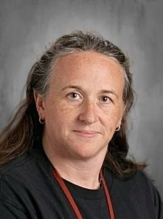 A woman with gray hair and light skin wearing a black top and a red lanyard, standing against a gray background.