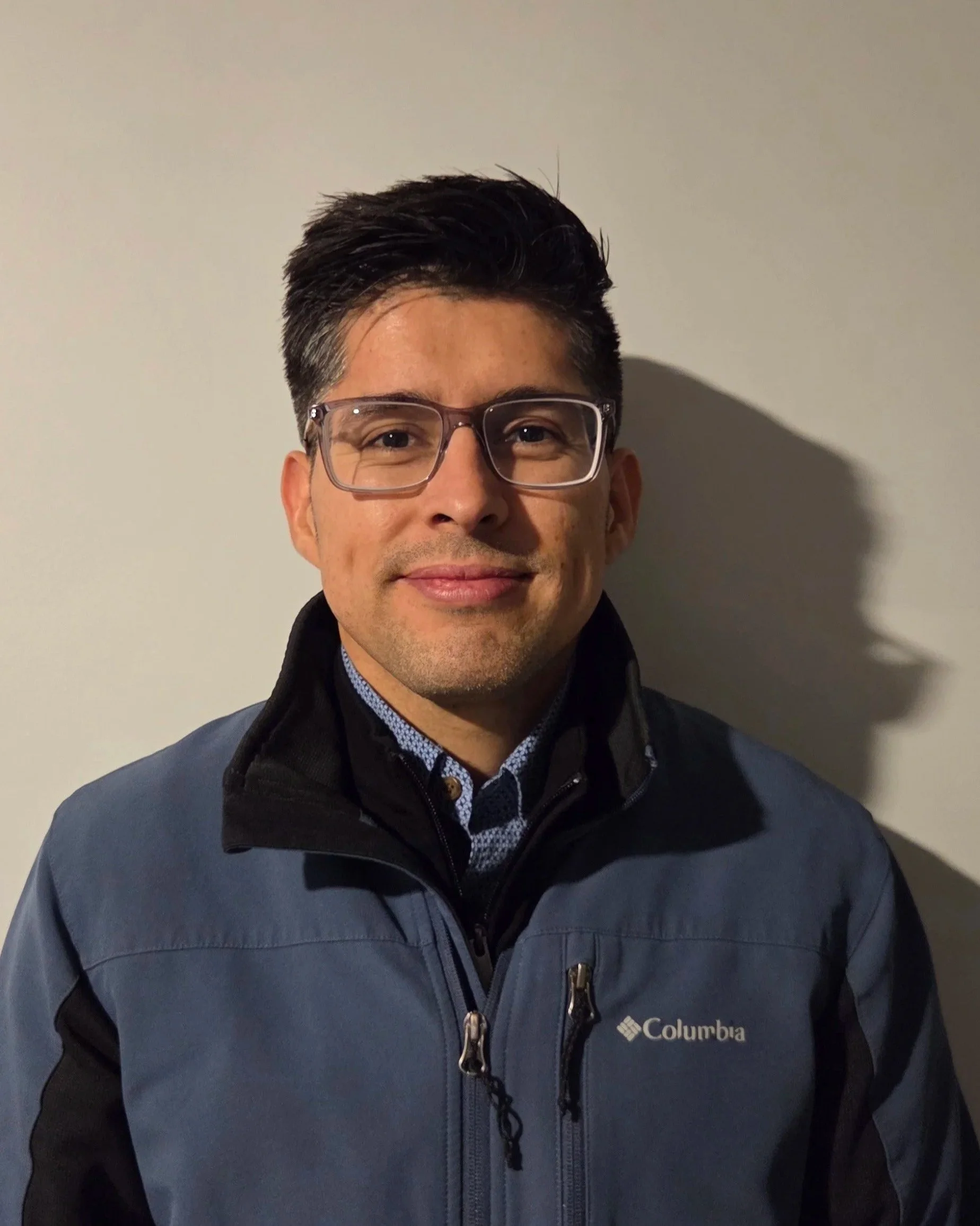 Headshot of a man wearing glasses and a Columbia jacket standing against a plain wall.