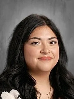 A young woman with long dark hair smiling, wearing a white patterned top, against a gray background.