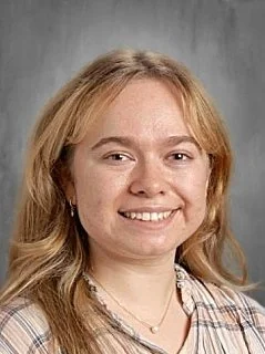 A woman with long, wavy blonde hair smiling at the camera, wearing a plaid shirt and a necklace.