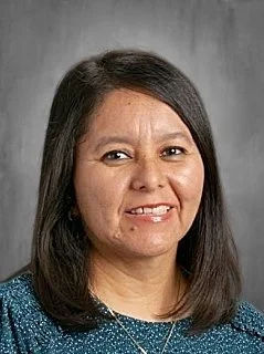 Portrait of a woman with shoulder-length dark hair, wearing a blue top and a necklace, smiling in front of a gray background.