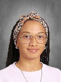 Portrait of a woman with glasses and braids, wearing a patterned headscarf and a white top, smiling against a gray background.