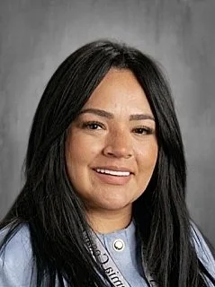 A woman with long black hair smiling, wearing a light blue shirt and standing against a gray background.
