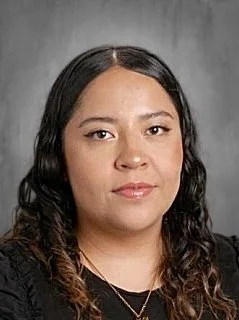 Portrait of a woman with long, wavy black hair and a neutral expression, wearing a black top and gold necklace against a gray background.