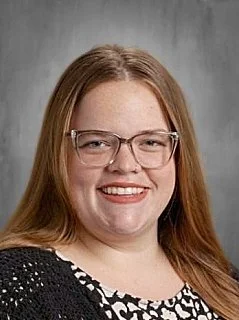 Portrait of a young woman with long red hair and glasses, smiling against a gray background.