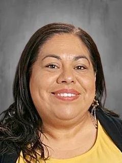 Close-up of a woman with dark hair, smiling, wearing a yellow top and earrings