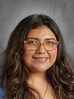 A woman with wavy dark hair wearing glasses and a gold necklace, smiling against a gray background.