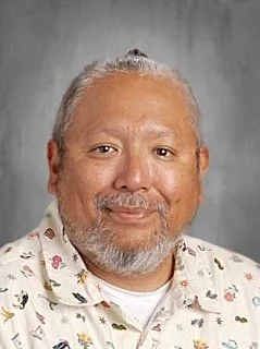 Portrait of a man with gray hair and beard, smiling, wearing a light-colored, patterned shirt, against a plain background.