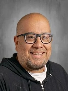 Close-up portrait of a bald man with glasses, smiling, wearing a black zip-up jacket against a gray background.