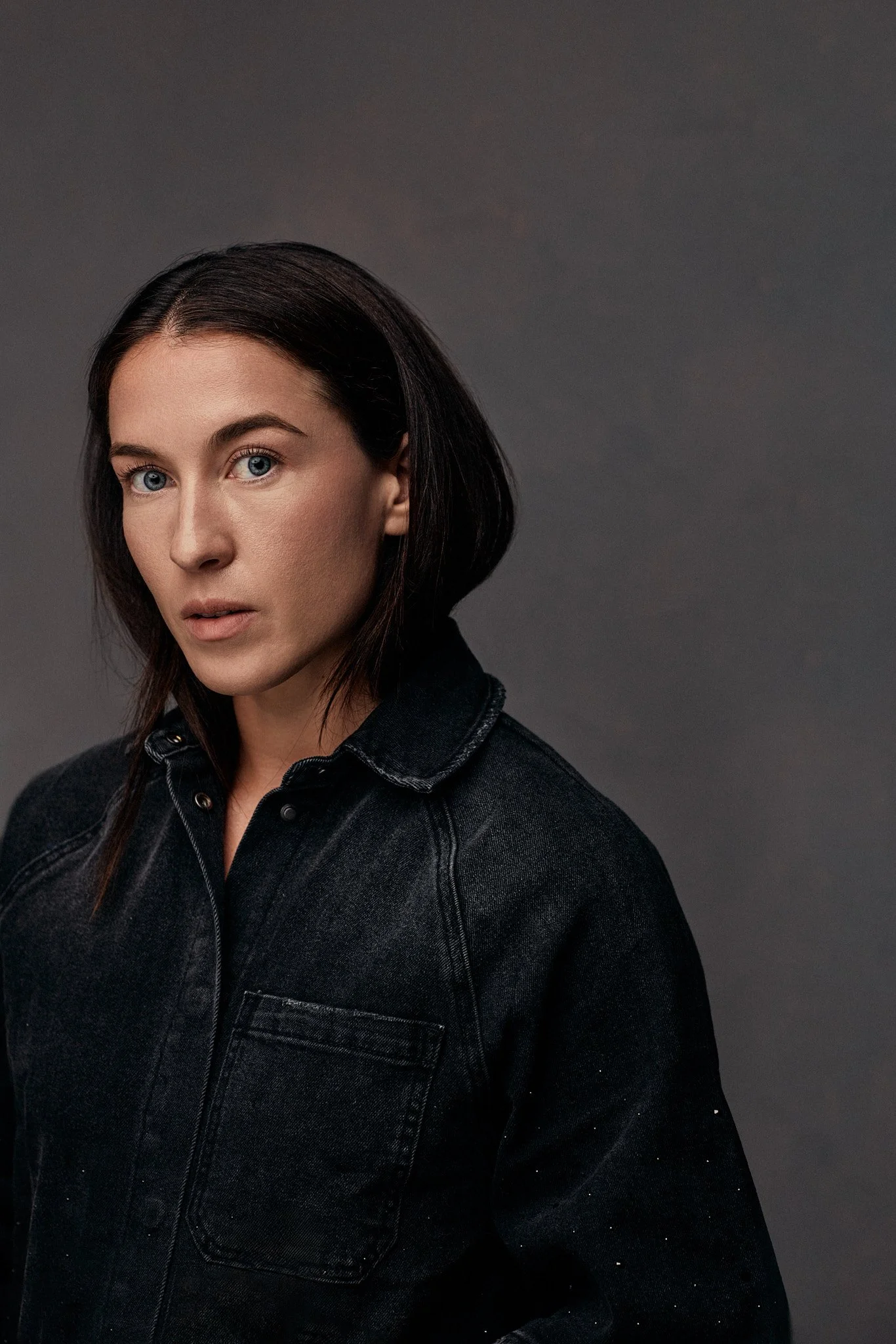 A woman with blue eyes and dark brown hair, wearing a black denim jacket, posing against a gray background.