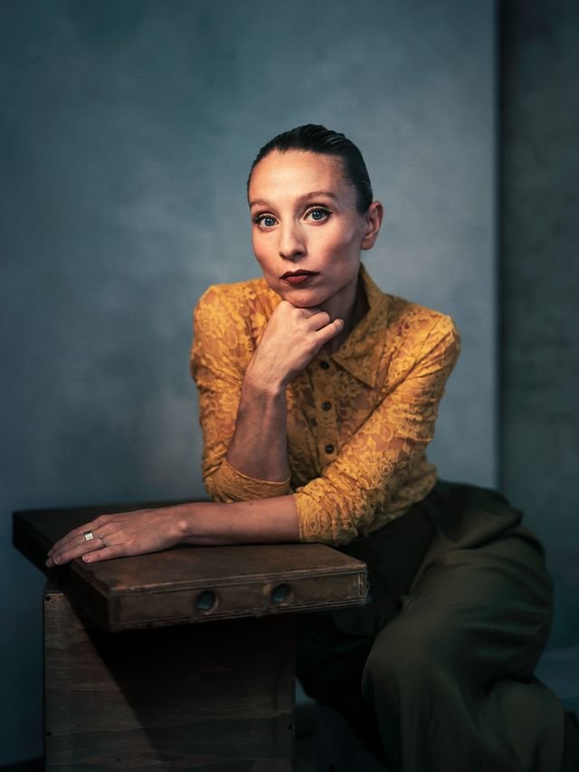 A woman with fair skin and brown hair pulled back, wearing a mustard yellow lace blouse, sitting at a dark wooden table with one arm resting on the table and her hand touching her chin, looking at the camera against a plain, muted gray background.