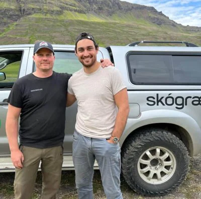 Two men standing in front of a silver truck with mountains in the background