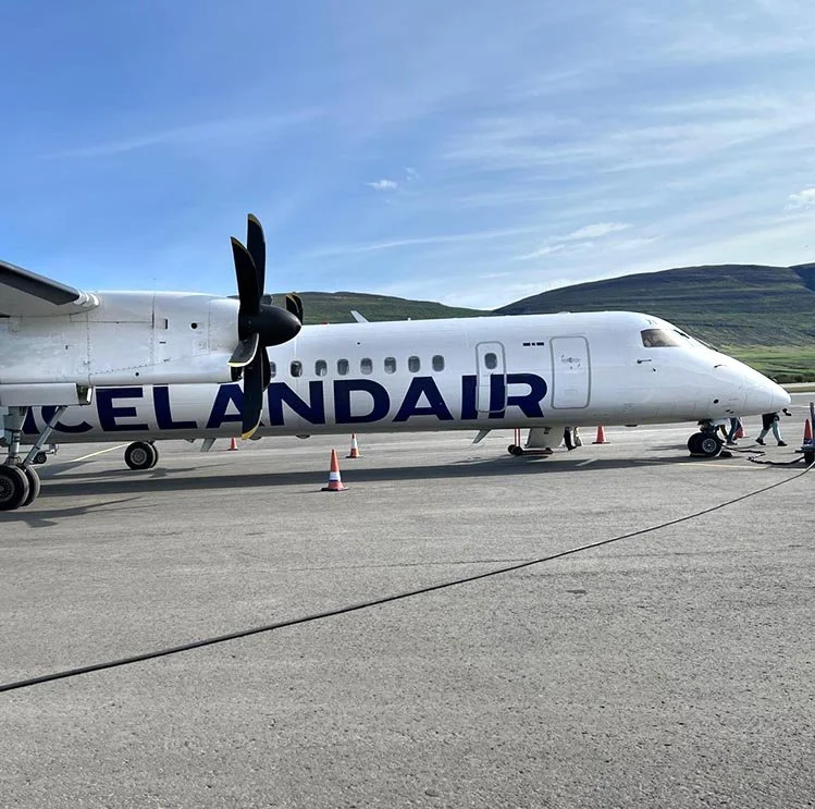 A white Icelandair airplane parked on the tarmac with orange cones around it, under a blue sky with some clouds and green hills in the background.