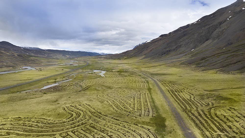Landscape of a valley with green fields, winding roads, and small waterways, surrounded by mountains under a cloudy sky.