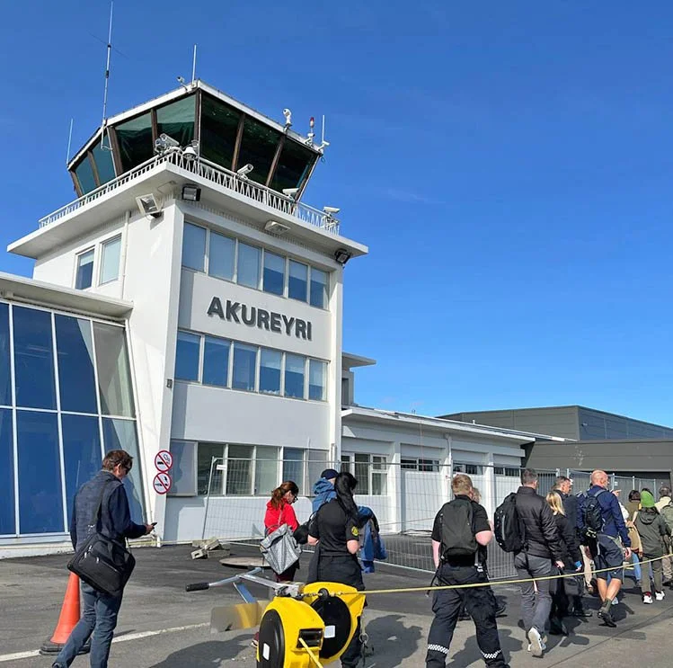 People walking near the Akureyri airport terminal, with the control tower visible against a clear blue sky.