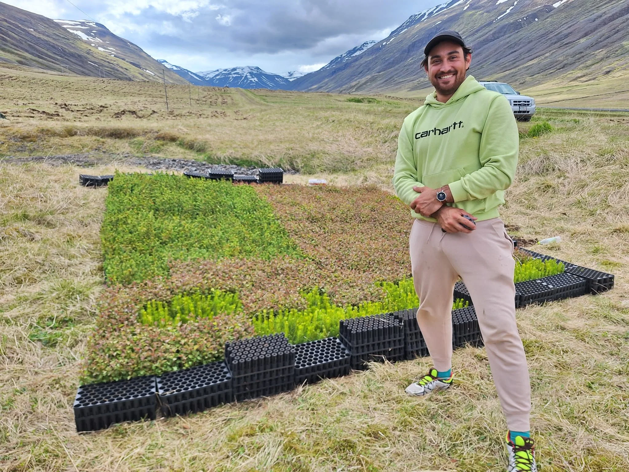 A man wearing a green hoodie, beige pants, and colorful shoes stands outdoors in front of trays of small plants, with mountains visible in the background.