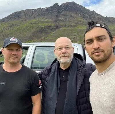 Three men taking a selfie outdoors in front of a mountain with a cloudy sky, standing next to a white vehicle.