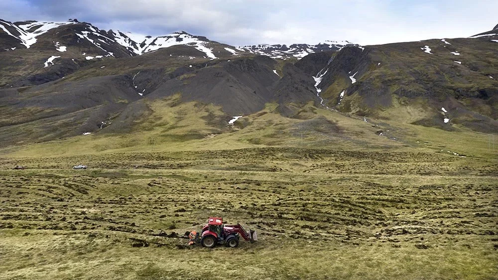 A red tractor working in a vast grassy field with rolling hills and mountains in the background, some snow patches on the mountains.
