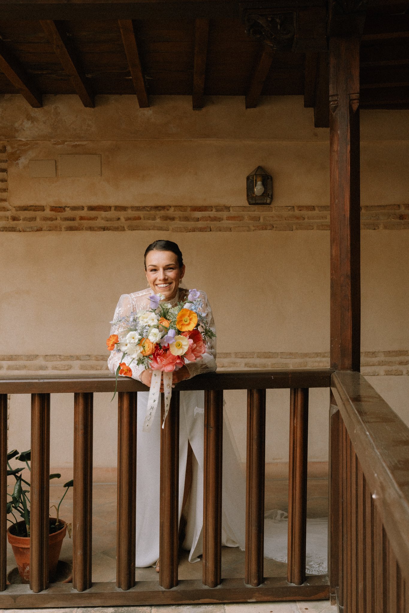 Une femme souriante en robe blanche, tenant un bouquet de fleurs colorées, se tient derrière une barrière en bois, dans un intérieur avec un mur en pierre et une lumière suspendue en forme de lanterne.