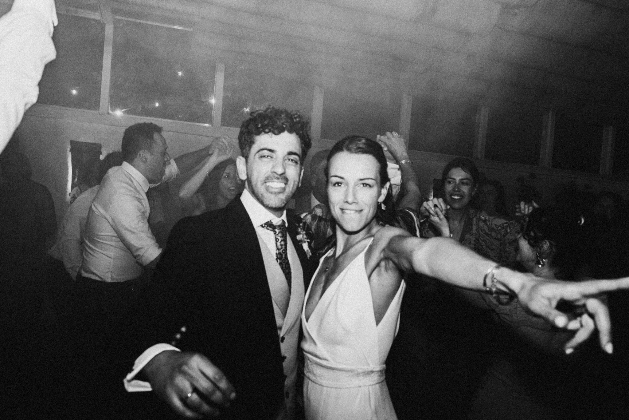 Une photo en noir et blanc de plusieurs personnes dansant à une fête ou un événement social, avec un couple au centre, le homme porte un costume et la femme une robe blanche, ils sourient et prennent un selfie.