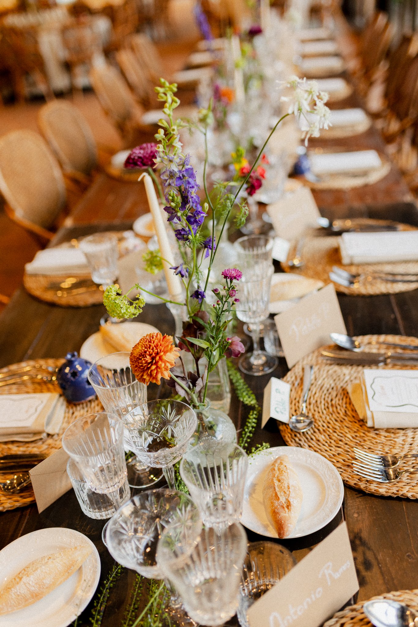 Une table de mariage décorée avec des fleurs, des assiettes, des verres en cristal, des baguettes, des serviettes pliées et des menus, dans une ambiance chaleureuse et élégante.